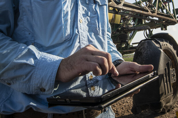 In an outdoor setting, in front of farm machinery, a man taps on a tablet device.