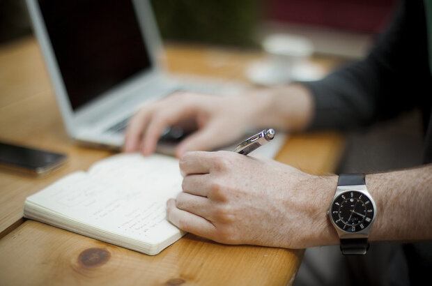 Someone writes in a notebook with laptop in the background