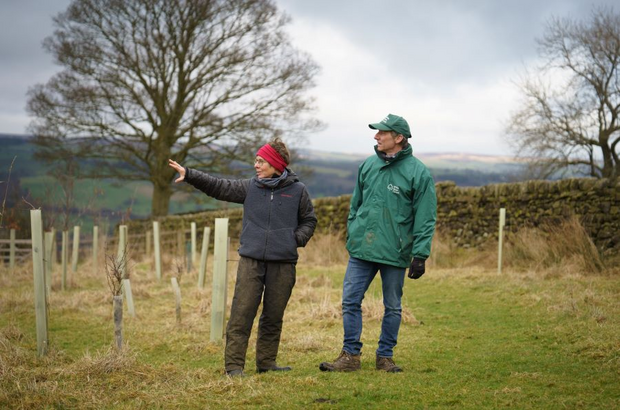 Two people talk in a countryside setting with tree in the background.