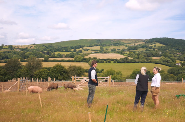 Rare breed pig farmers Sarah and Byron Odell are visited on their Shropshire farm by adviser Karen Fisher from the Soil Association. They stand in a field with pigs in the background.