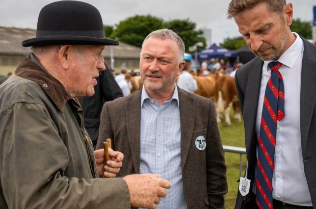 Steve Reed, Environment Secretary, stands outdoors at a livestock show talking with two men. He is in the centre, wearing a brown jacket and light blue shirt, listening attentively. On his left, an older man in a dark coat and black bowler hat gestures while speaking. On his right, a suited man with a red and blue striped tie looks down thoughtfully. Cattle and people are visible in the background