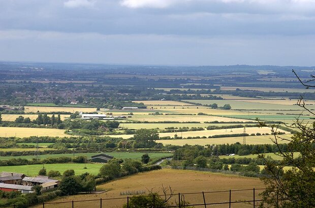 A wide view of the English countryside with patchwork fields, hedgerows, farm buildings, and clusters of trees under a cloudy sky. Rolling hills stretch into the distance, with a village partially visible on the horizon.