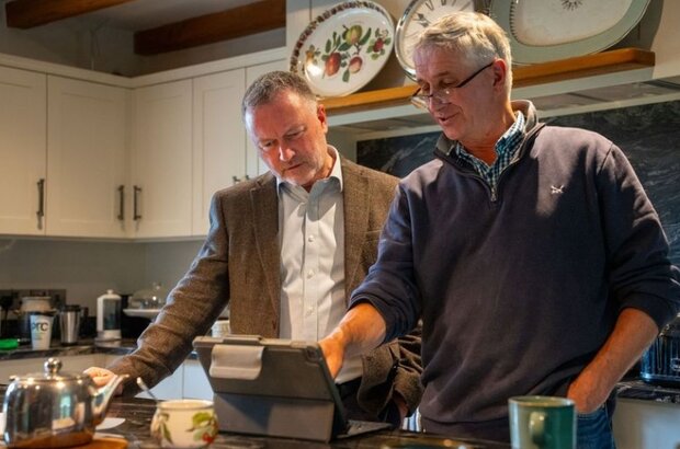 Alt text: Secretary of State Steve Reed stands beside farmer Mike Roberts in a farmhouse kitchen. They are looking at a tablet screen together, with mugs and a teapot on the counter in front of them. Decorative plates are displayed on a shelf above. The atmosphere appears informal and focused.