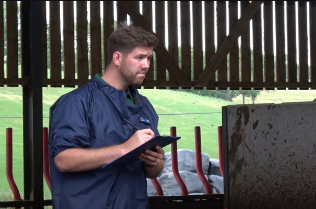 A vet in waterproof overalls carries out a farm visit. He stands with clipboard and pen in hand.