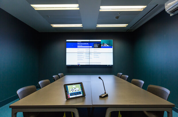 An image of a user research lab in government. The one pictured is GDS User Research Lab, a long table and two screens with a microphone.