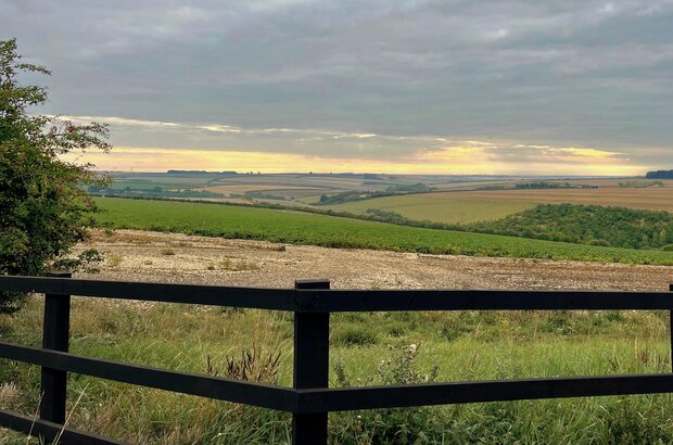 Landscape at the end of the day. Grey skies are pierced with yellow light, a patchwork of fields in the distance and a fence in the foreground.
