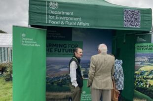 Team member in green gilet talking to a man in tweed in front of the Defra stand at an event.