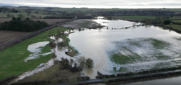 Ariel view of water across the landscape in sweeping curves, with patches of grass, bare trees, and agricultural land visible beneath the floodwater.