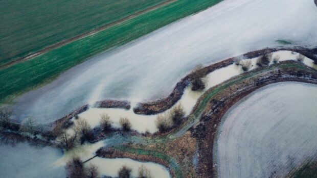 An aerial view of a meandering river channel surrounded by flooded fields. Water spreads across the landscape in sweeping curves, with patches of grass, bare trees, and agricultural land visible beneath the floodwater