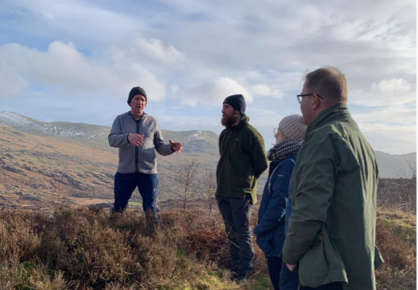 A group of 4 people stand on open land. One is talking to the group who stand listening under cloudy skies.