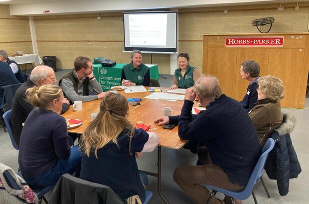A group of people sit around a rectangular wooden table in a community hall, engaged in discussion. Papers, notebooks, and mugs are spread across the table. A presentation screen at the front of the room displays a slide, and a banner for the Department for Environment, Food & Rural Affairs is visible on a table nearby. A podium with a “Hobbs Parker” sign stands to the right.