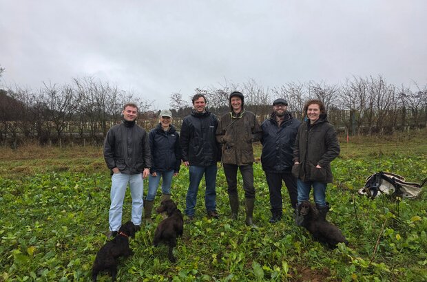 Six people stand in a green, leafy field on an overcast day, with several small black dogs sitting or standing at their feet. Behind them is a line of bare shrubs and trees.