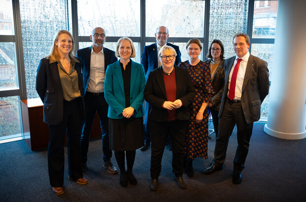 A group of eight people stand together in a well‑lit indoor space in front of large floor‑to‑ceiling windows. They are dressed in business or smart‑casual clothing, arranged in two rows with five individuals in the back and three in the front. Behind them, the windows reveal an outdoor view with trees and nearby buildings. The room has dark carpeting and a modern interior with a column visible on the right side of the image. Environment Secretary Emma Reynolds is pictured with Farming Minister Dame Angela Eagle.