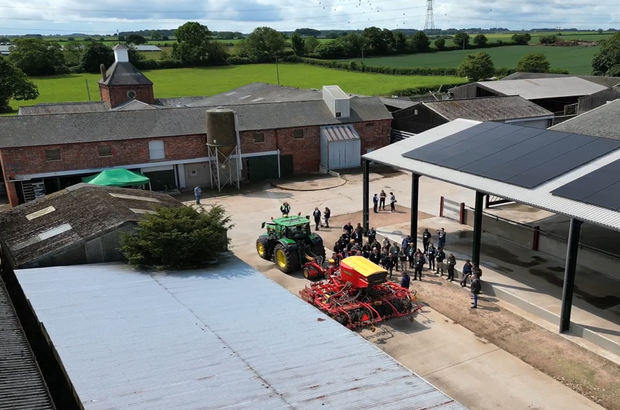 A high, wide-angle view of a farmyard with several brick and metal-roofed buildings surrounded by green fields. In the center of the yard, a green tractor is attached to a large red and yellow agricultural implement. A group of people stands gathered around the machinery, appearing to observe or take part in a demonstration. Some structures have solar panels on their roofs, and the weather is bright with scattered clouds.