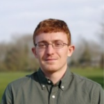 Photograph of a person with short red hair wearing a dark green button-up shirt standing outdoors with blurred trees and sky in the background.