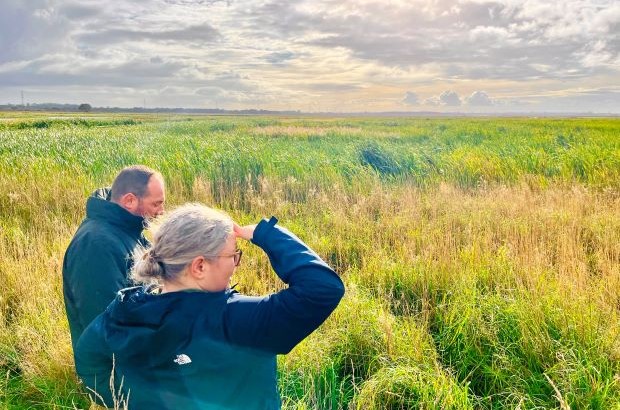Photograph showing two people standing in a grassy field under a cloudy sky, with one person shielding their eyes while looking into the distance. The scene highlights a natural landscape with tall green and yellow grasses extending toward the horizon.