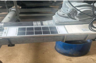 An irrigation controller mounted on a metal beam, featuring a rectangular panel with wiring and control components above it, installed as part of a farm irrigation system