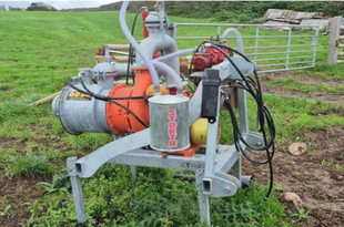 A slurry chopper pump mounted on a metal frame in a grassy field, with an orange cylindrical tank, a silver motor, white hoses, and black electrical cables connected to the unit