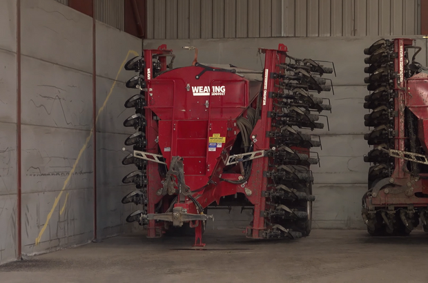 A red Weaving direct drill with folded side frames and multiple rows of metal seed tines is parked indoors against a concrete wall inside a large farm shed.