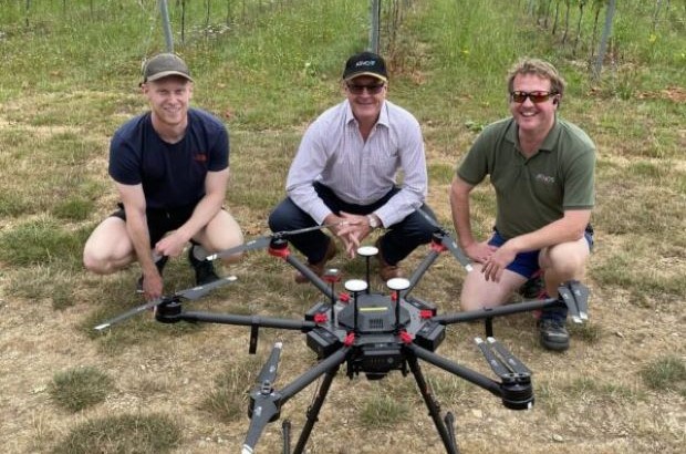 Three people crouched down in a vineyard with a drone in front of them on the ground.