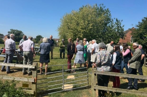 A group of people gathered in a grassy field behind a farm gate during a community outdoor event, with trees and houses in the background on a sunny day.